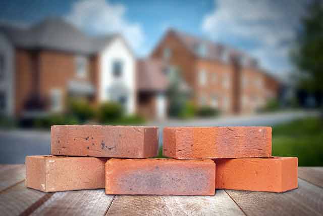 Five Brown/Red House Bricks layed on a wooden table in front of a housing estate