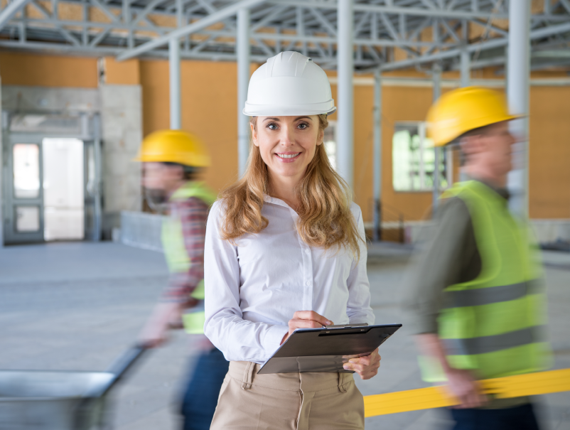 Construction Inclusion Coalition Lady with a white hard hat and clipboard with a blurred construction background with two workmen
