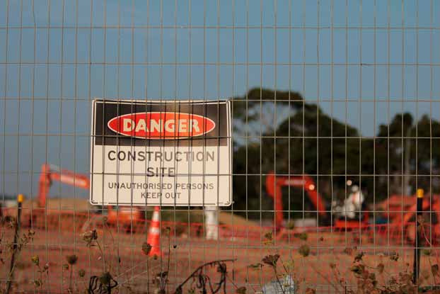 A large sign reading Danger Construction site unauthorised persons keep out taken through a fence showing construction site machinery