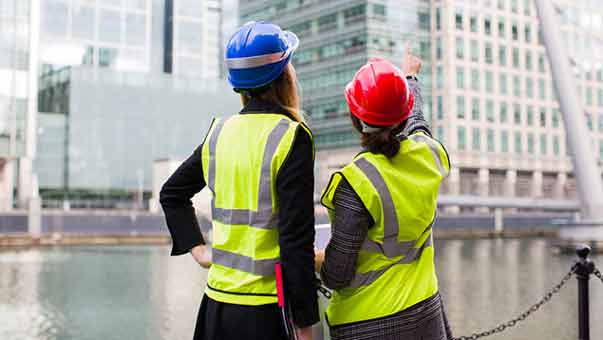 Two construction women with high vis and hard hats in a city in front of skyscrapers with one woman pointing at a skyscraper.