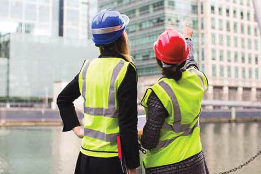 Two construction women with high vis and hard hats in a city in front of skyscrapers with one woman pointing at a skyscraper.