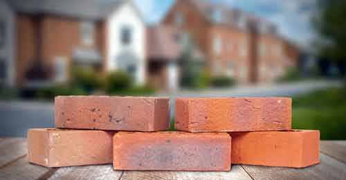 Five Brown/Red House Bricks layed on a wooden table in front of a housing estate