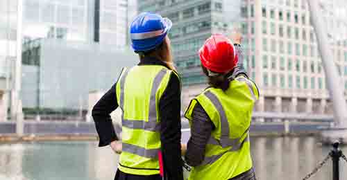 Two construction women with high vis and hard hats in a city in front of skyscrapers with one woman pointing at a skyscraper.