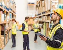 Warehouse worker with a clipboard shown within a warehouse full of boxes and packages with other warehouse workers